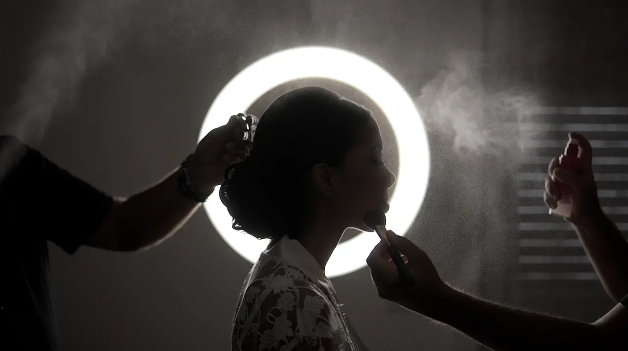 La sposa in controluce davanti a una luce anulare, tra mani che sistemano trucco e capelli