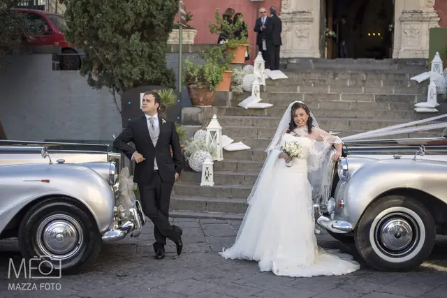 Gli sposi in posa tra due auto d'epoca davanti alla chiesa